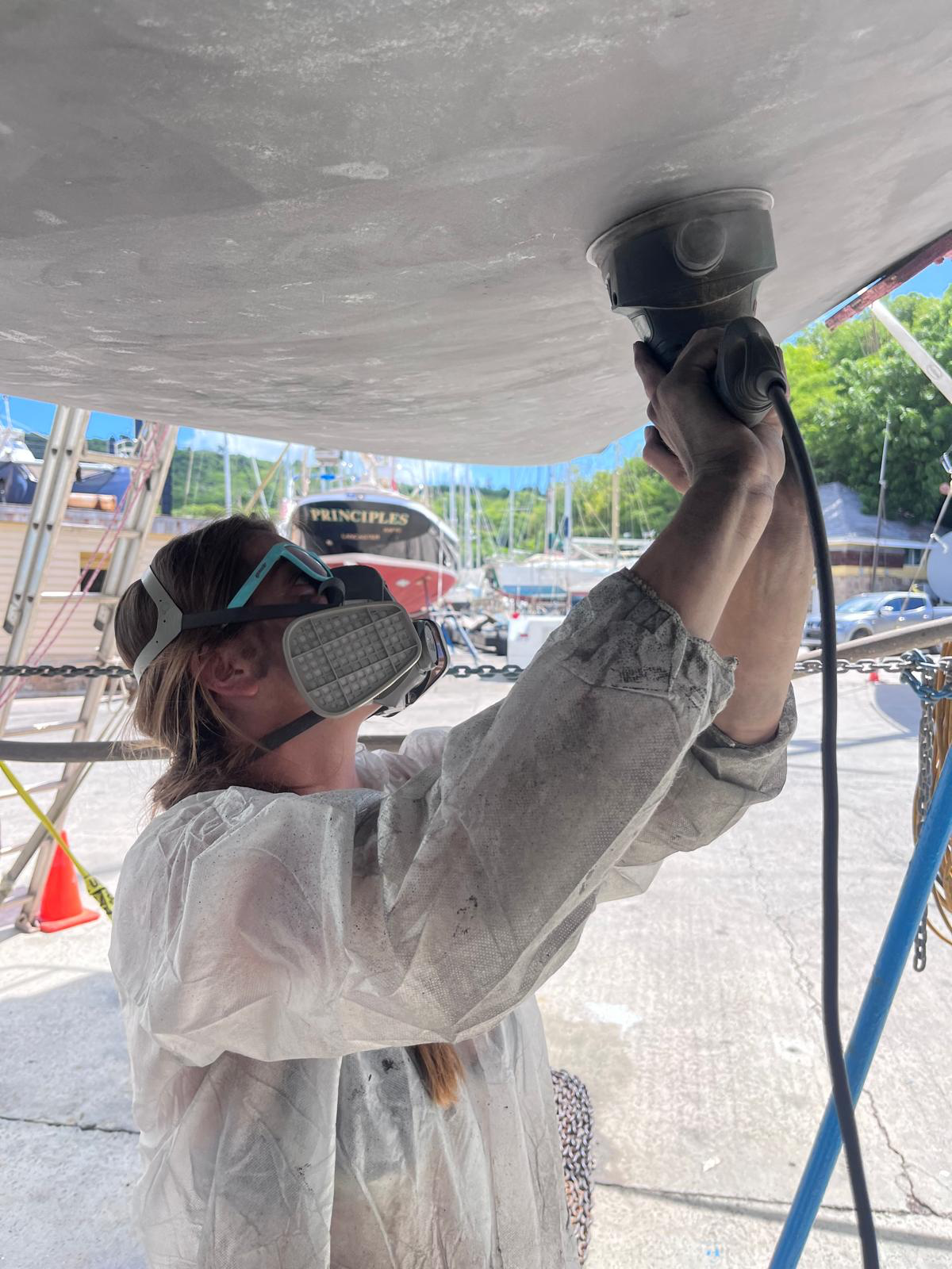 A Maritime Gal in a respirator sanding the bottom of a hauled-out sailboat in the boatyard